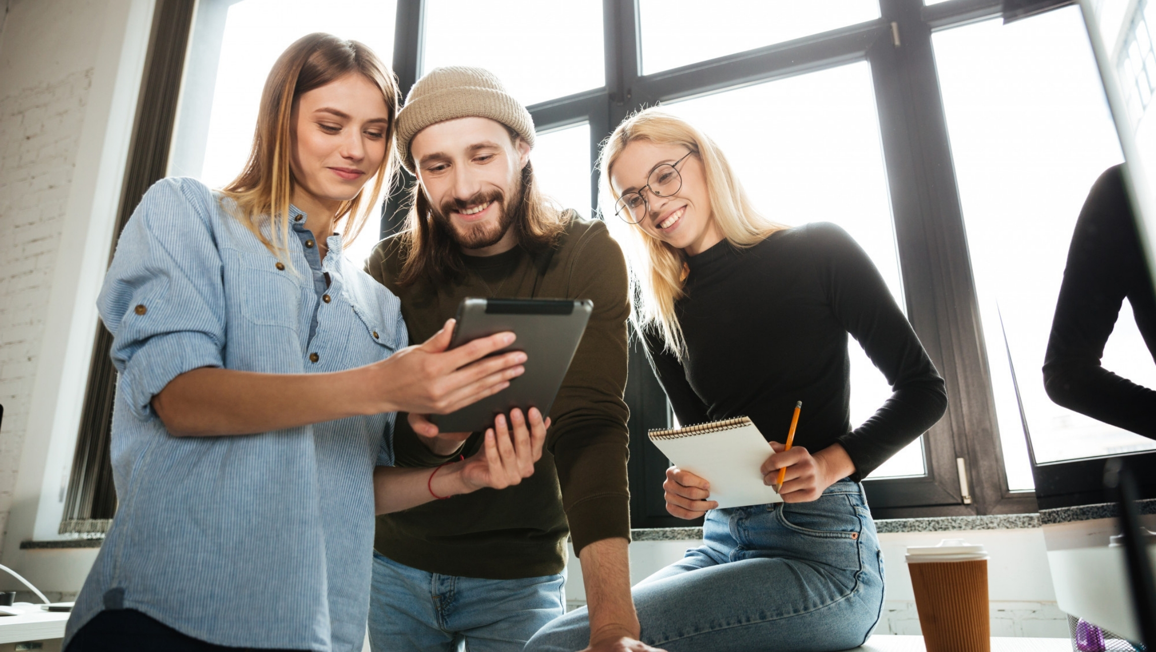 Dwie kobiety i mężczyzna w biurze patrzą z uśmiechem na ekran tabletu; jedna z kobiet trzyma w rękach notatnik i ołówek, przygotowując się do robienia notatek.