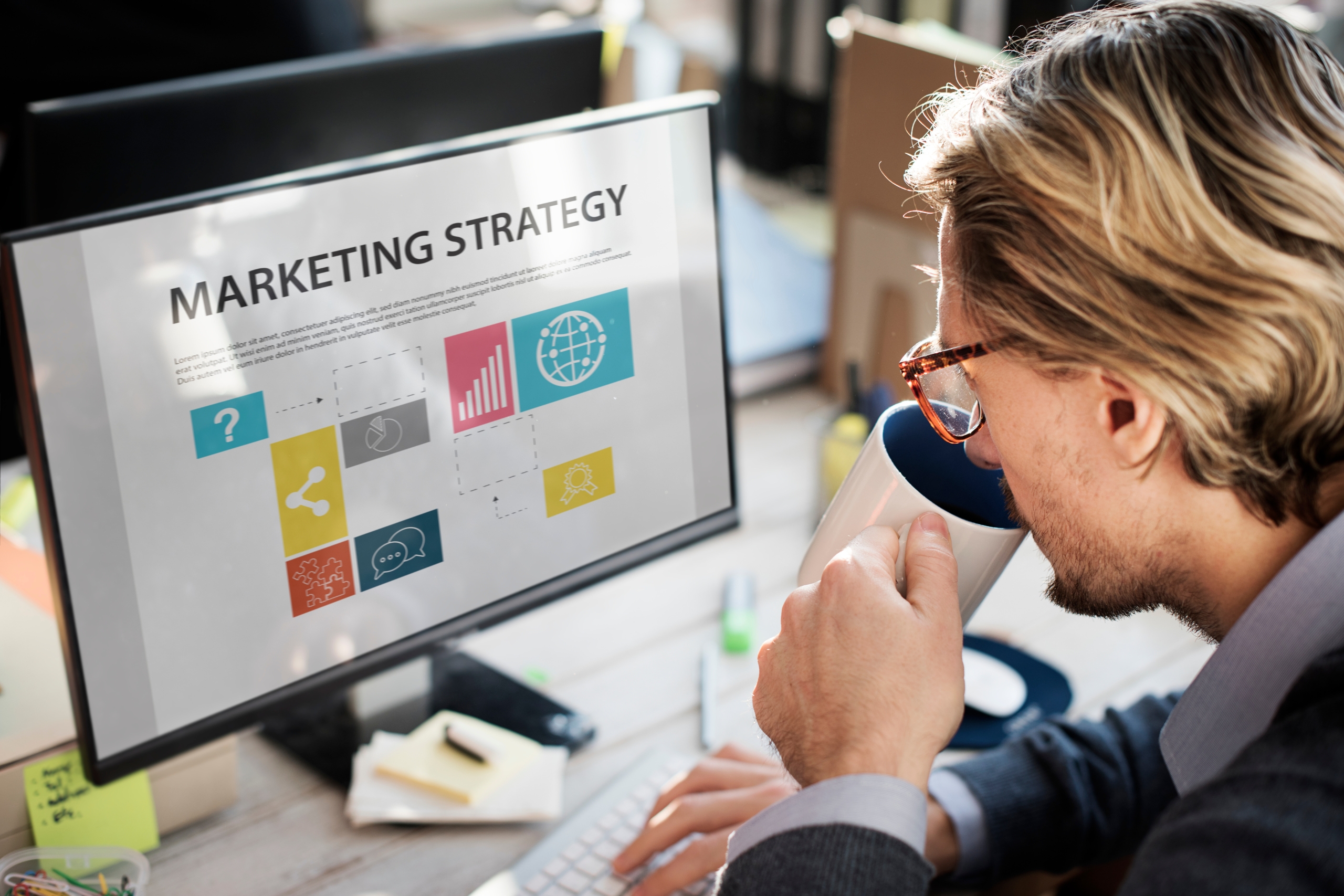 Mężczyzna w okularach pijący z kubka i patrzący na monitor komputera z wyświetloną prezentacją „MARKETING STRATEGY” zawierającą kolorowe ikony i wykresy.